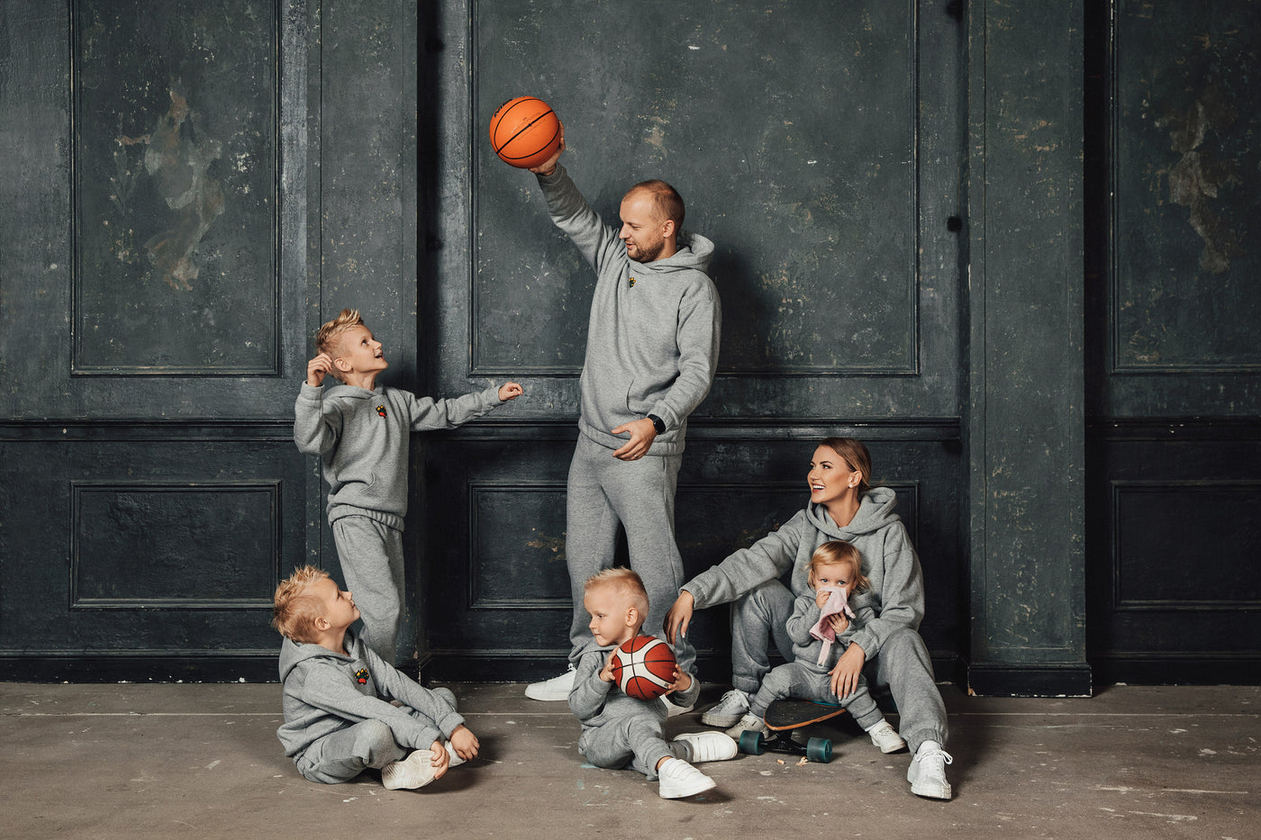 Tėtis ir vaikai žaidžia su krepšinio kamuoliu apsirengę pilkais laisvalaikio kostiumais